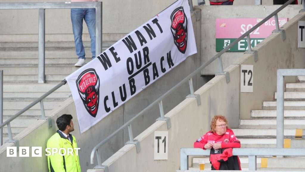 A Salford Red Devils banner displaying the message "we want our club back" at a Super League fixture against Wigan Warriors this season
