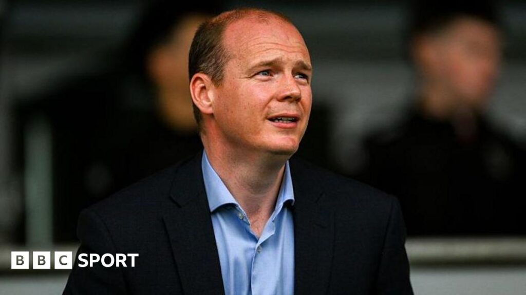 Gordon Lyons is looking to the right of the screen. He is wearing a light blue shirt and black blazer. He is slightly balding but has dark hair.